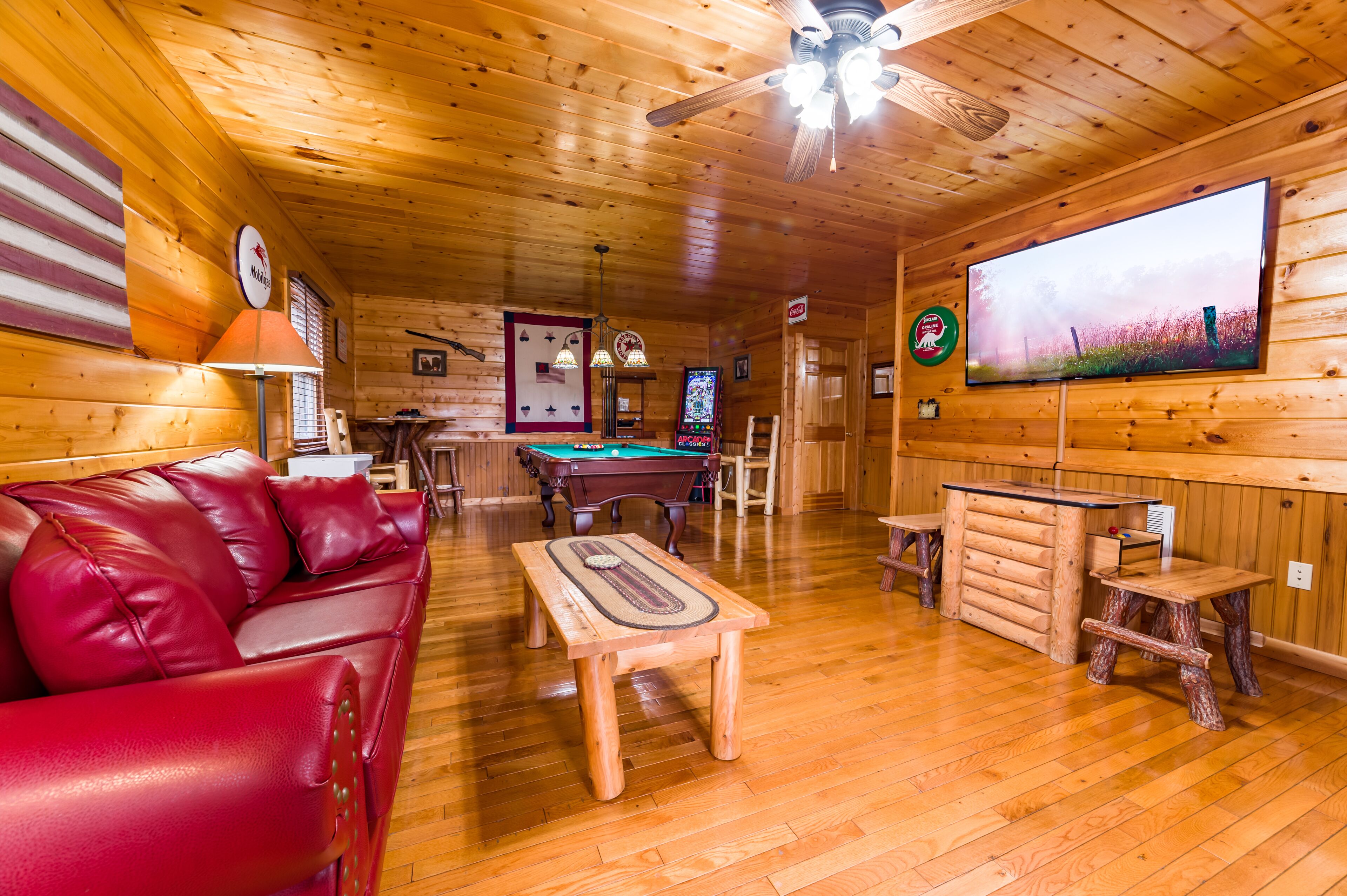 Un acogedor interior de cabaña con mesa de billar, sofá de cuero rojo, mesas de madera y un televisor grande en la pared.