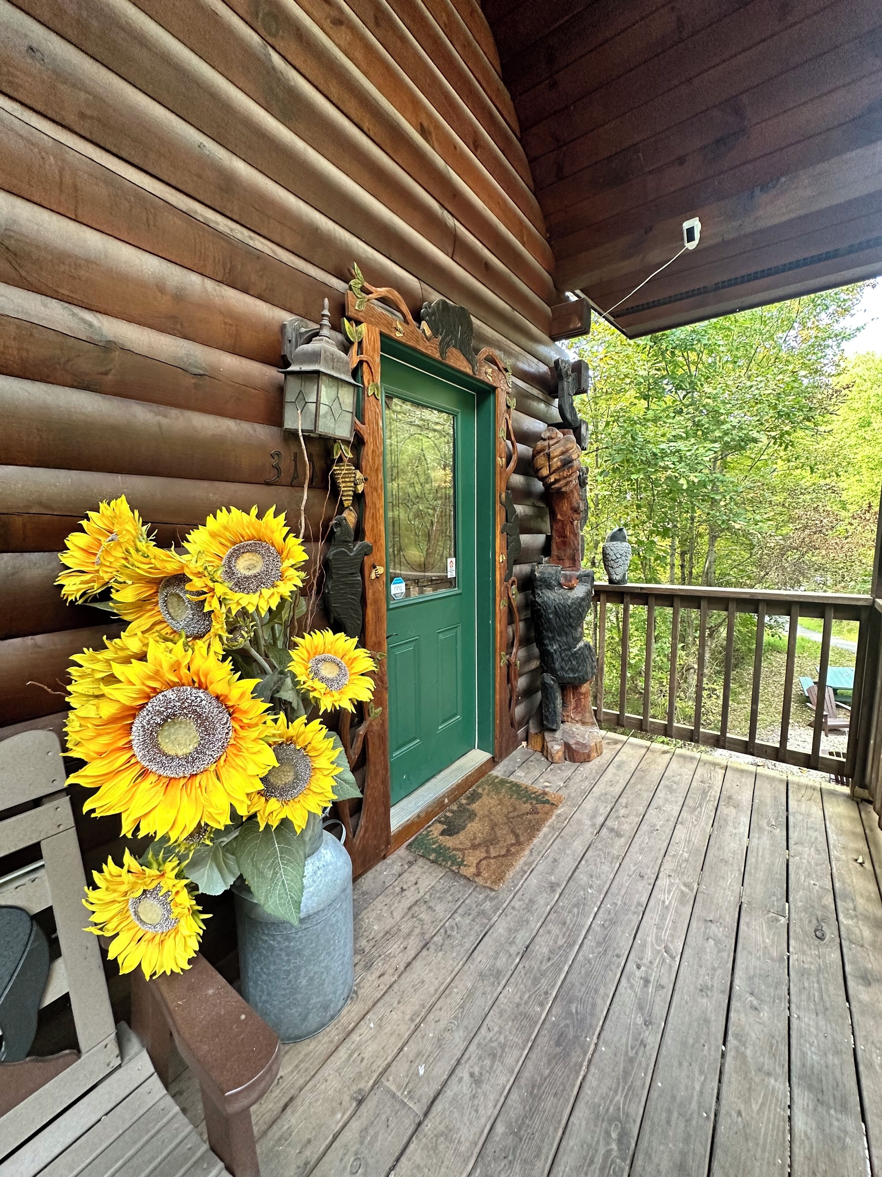 Une cabane en bois avec une porte verte, une véranda dotée d’une balustrade et un grand vase rempli de tournesols.