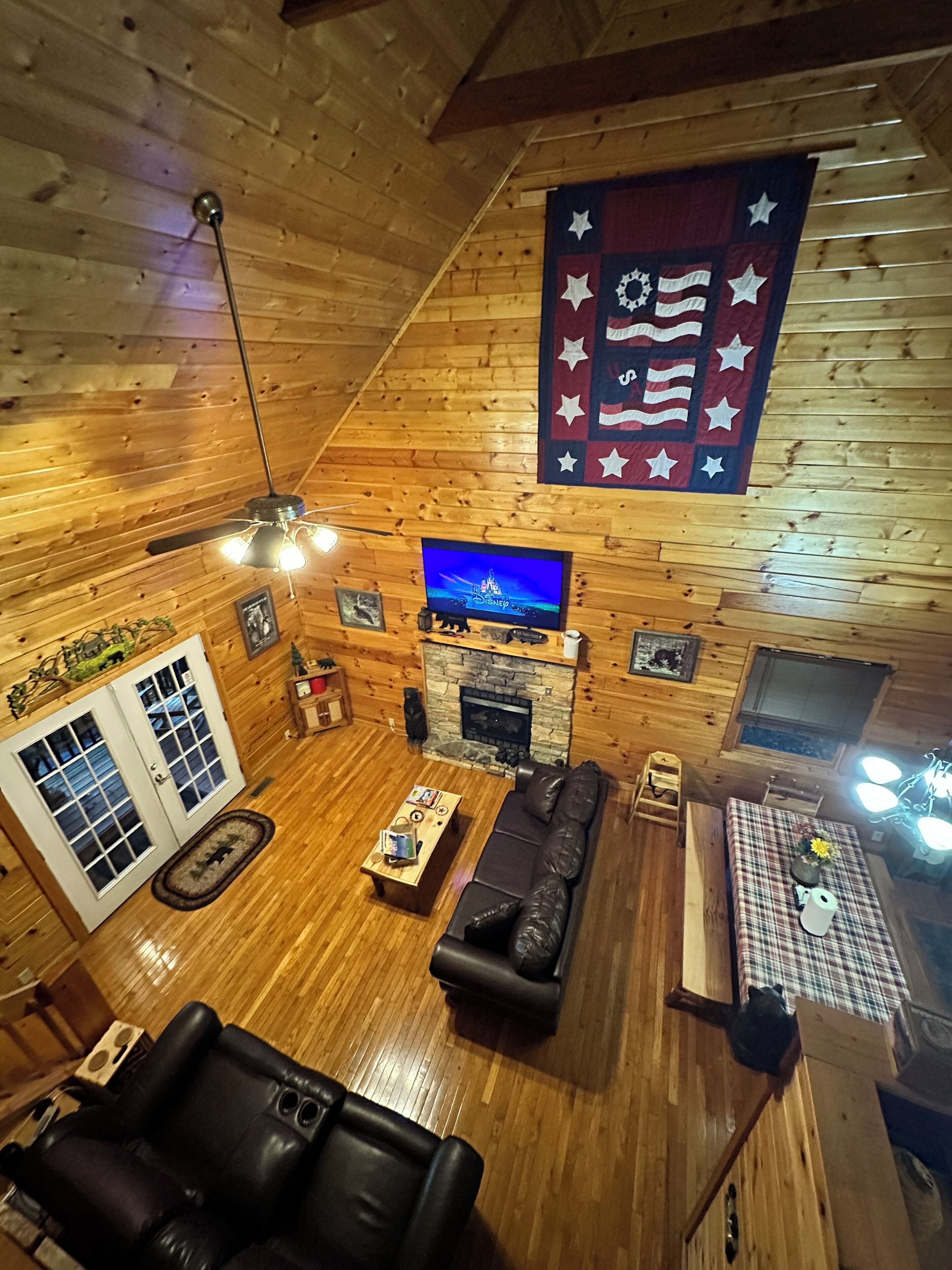 Interior acolhedor de uma cabana, com decoração em madeira, lareira de pedra, televisão e uma bandeira na parede.