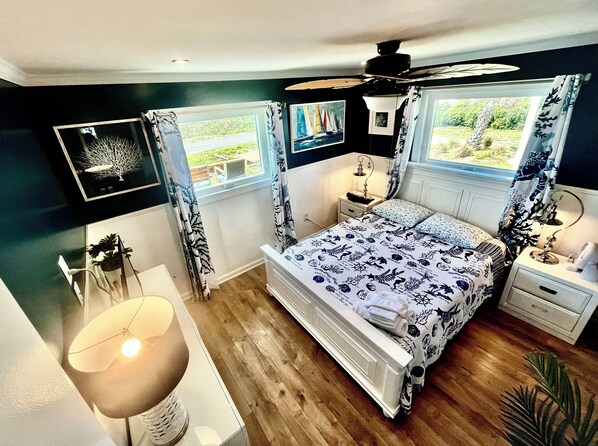 Un dormitorio con cama, mesitas de noche, lámpara, ventilador de techo y ventana con cortinas.