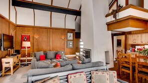 A modern living room with a grey sectional sofa, wooden accents, a TV, and a dining area with wooden chairs and a table.
