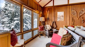 Une pièce chaleureuse avec de grandes fenêtres, une cheminée, un canapé et des paires de skis appuyés contre le mur.