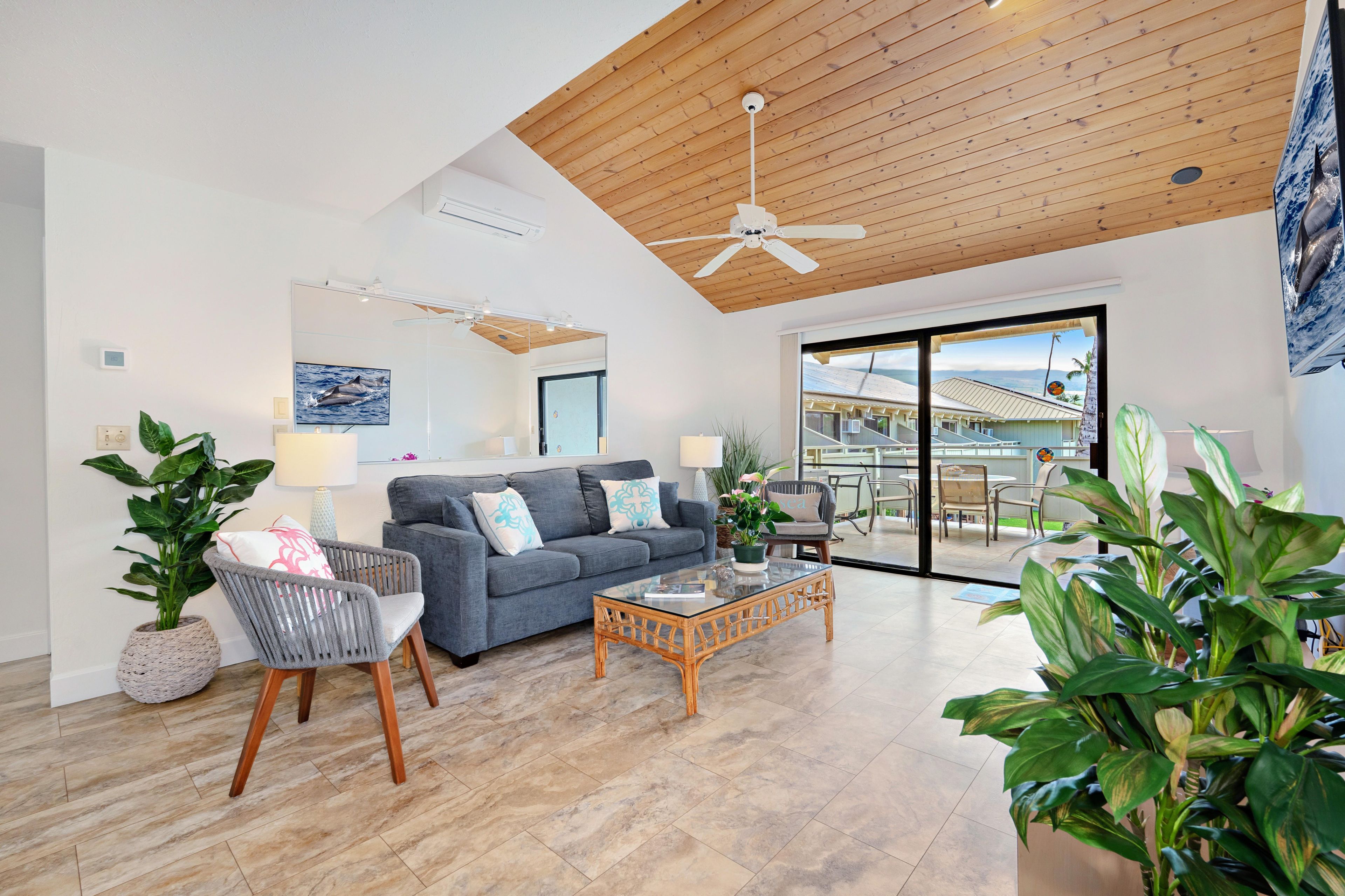 A spacious living room with a grey sofa, wicker chairs, a glass coffee table, and large windows offering a view of the outside.