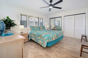 Une chambre à coucher comprenant un lit, un ventilateur de plafond, une fenêtre avec des stores, un miroir et une porte.