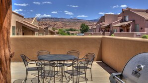 Eine Terrasse mit rundem Tisch und Stühlen, einem Grill und Blick auf ein Gebirge.