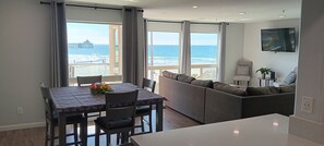 Un salon moderne doté d’une grande fenêtre donnant sur l’océan, d’un canapé, d’une table à manger avec des chaises et d’un téléviseur à écran plat fixé au mur.