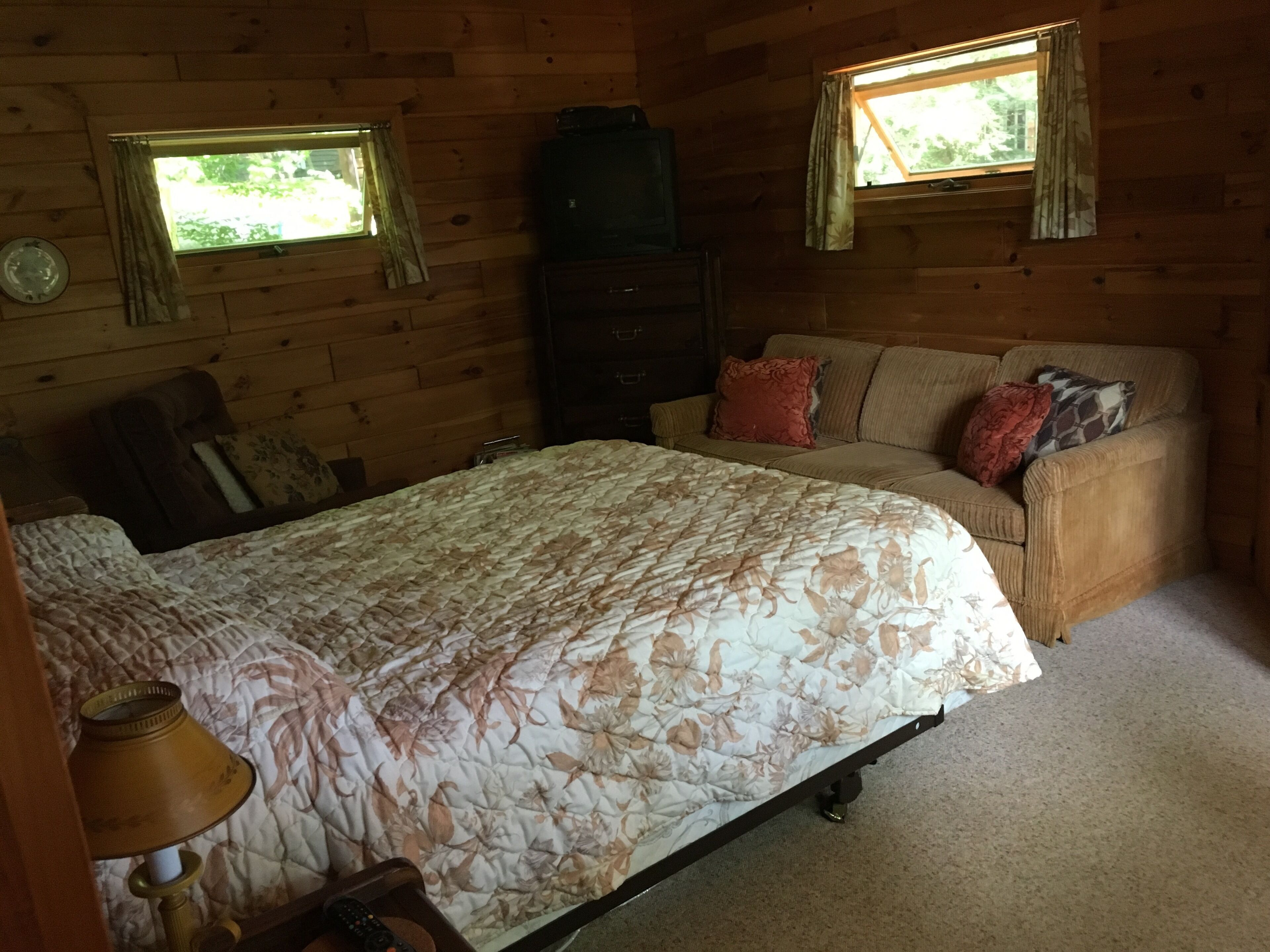 Una habitación de cabaña de madera con una cama, un sofá, un aparador y dos ventanas.