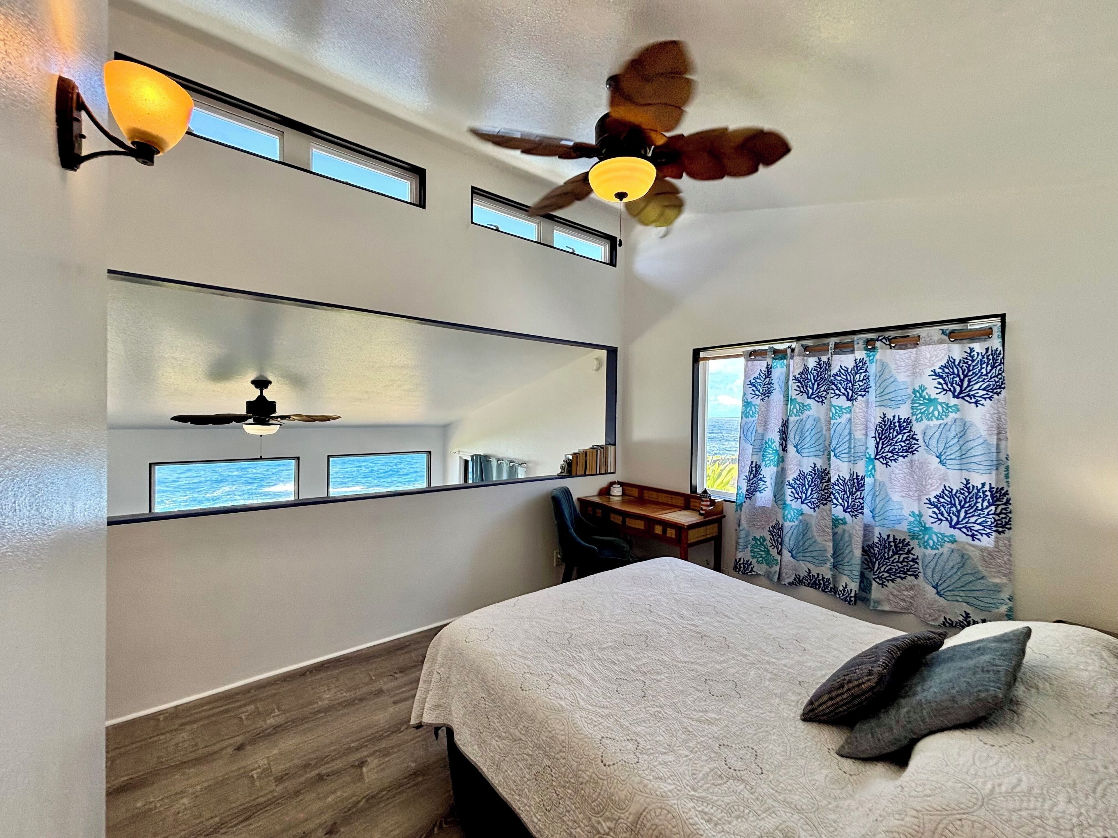A bedroom with a bed, a desk, a chair, a ceiling fan, a window with a view, and a mirror.
