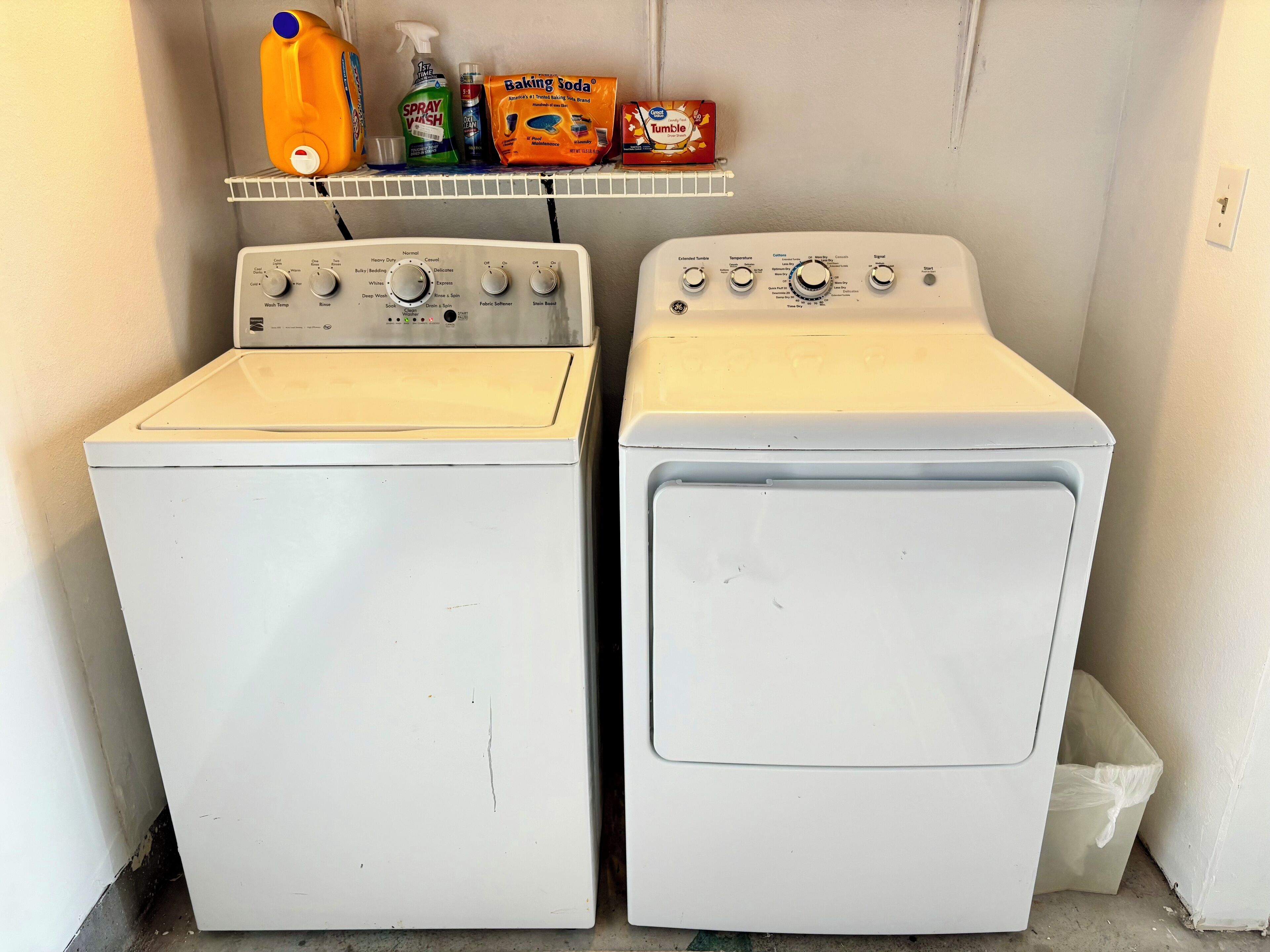 Two white front-loading washing machines with control panels, a shelf above containing cleaning products, and a trash bin to the right.