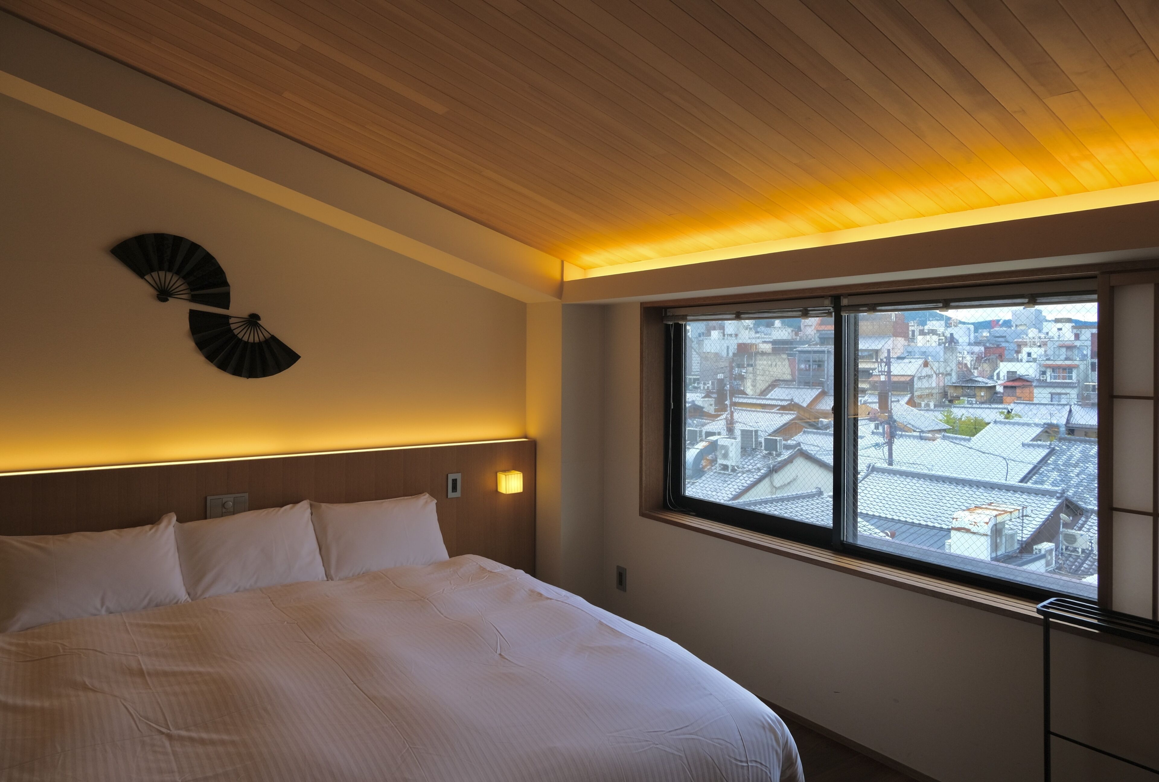A hotel room with a large bed, a wooden ceiling, a wall-mounted fan, and a window with a city view.