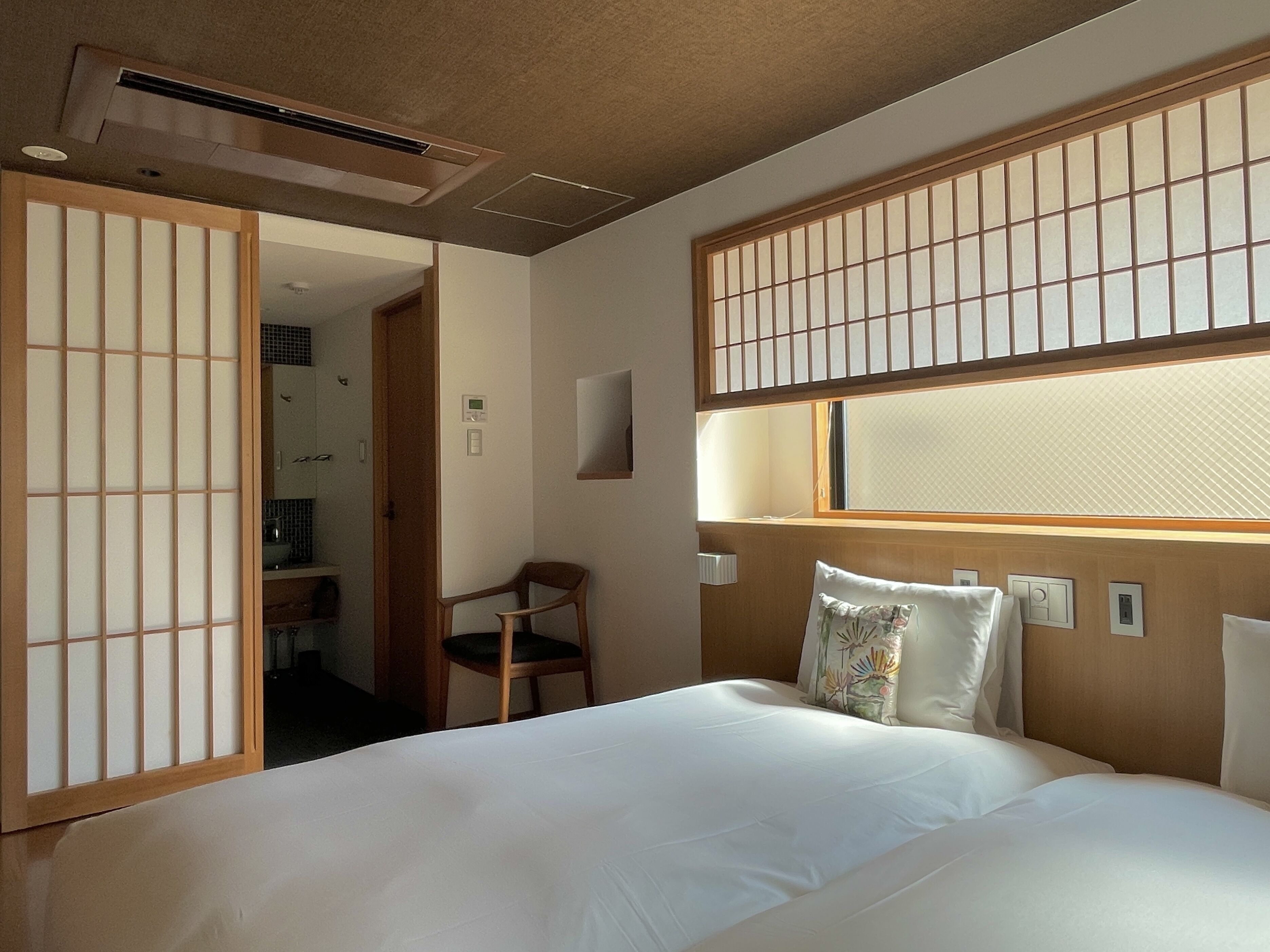 A hotel room with a bed, a chair, a window with a view, and a traditional Japanese shoji screen.