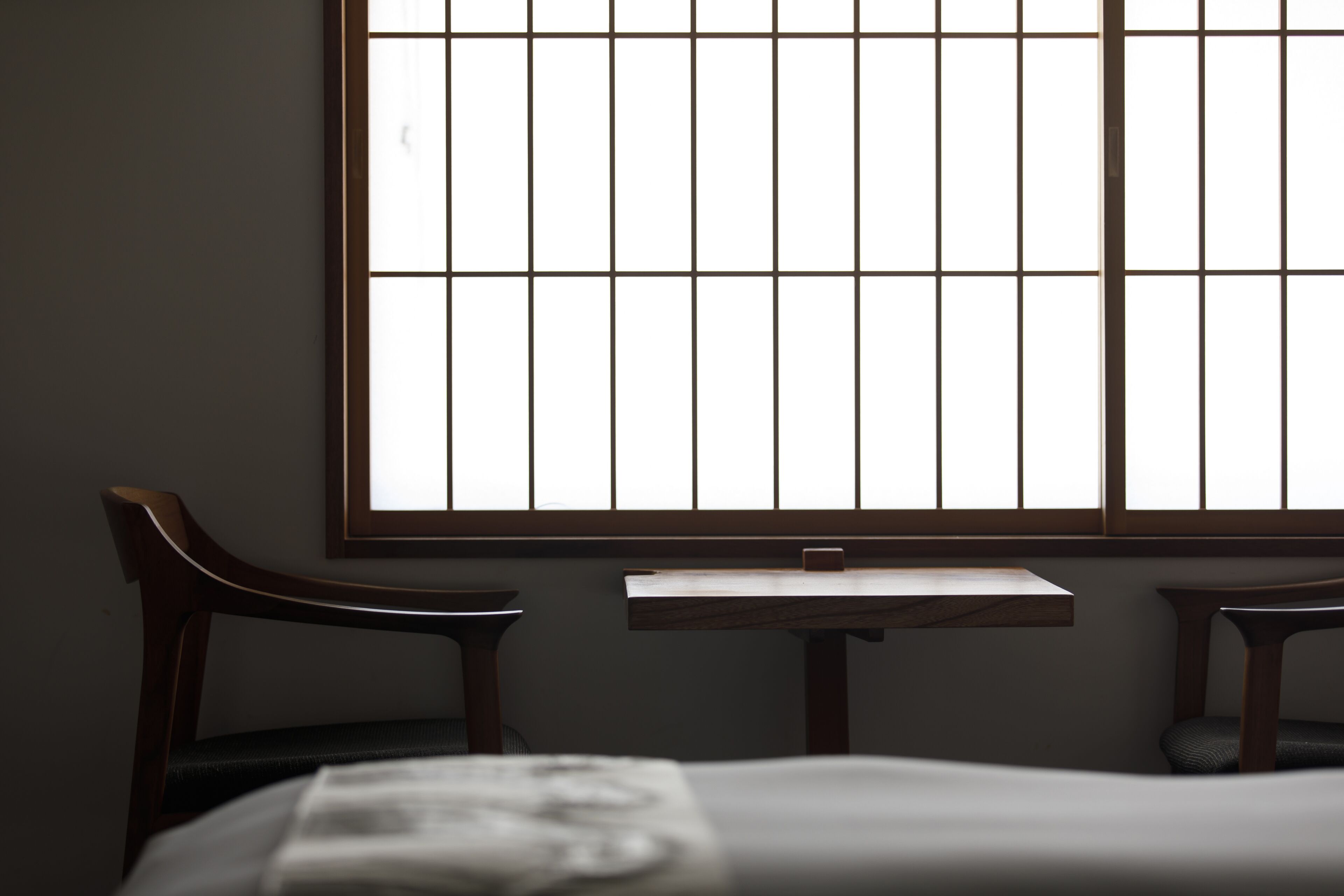 A room with a large window, two wooden chairs, a small table, and a bed.