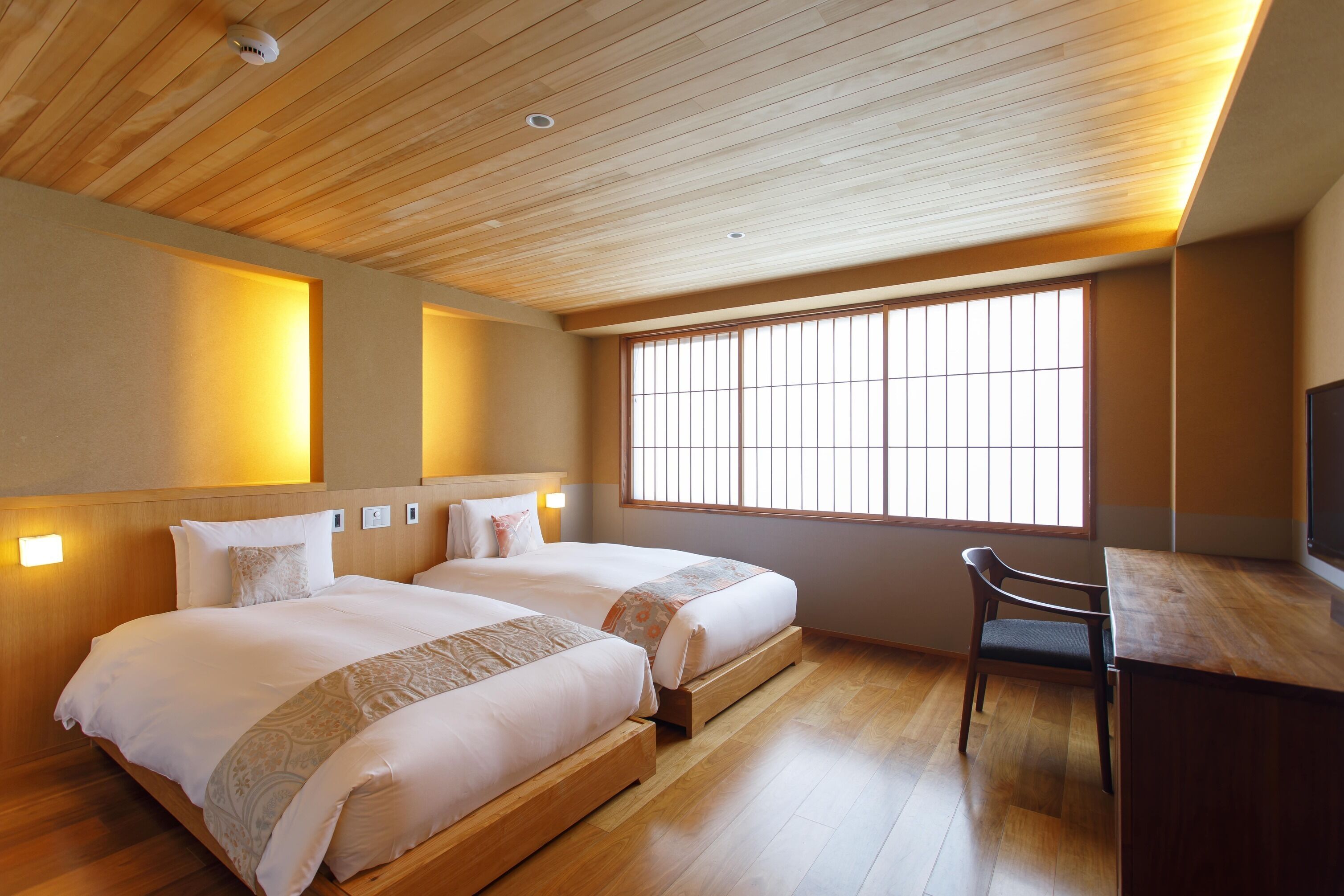 Ein Hotelzimmer mit zwei Betten, einem Schreibtisch, einem Stuhl, einem großen Fenster und einer Holzbalkendecke.
