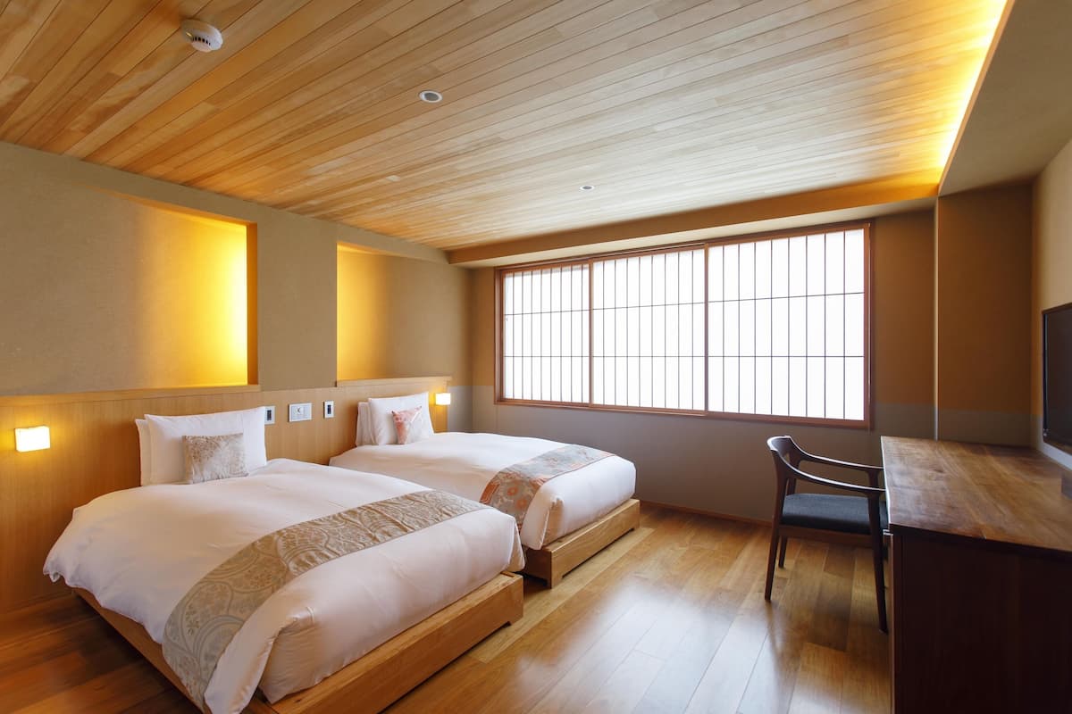 A hotel room with two beds, a desk, a chair, a large window, and wooden ceiling.