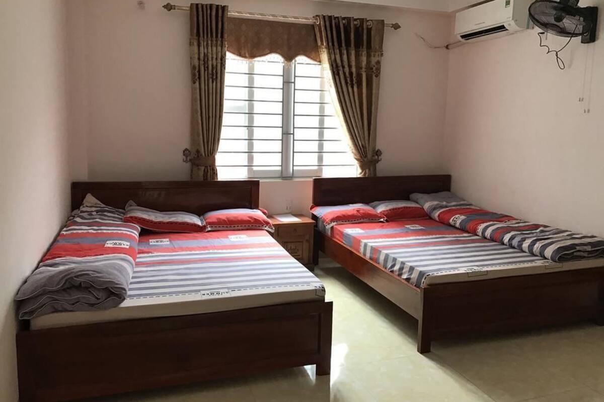 Une chambre avec deux lits en bois, une fenêtre avec des rideaux, un climatiseur et un ventilateur fixé au mur.