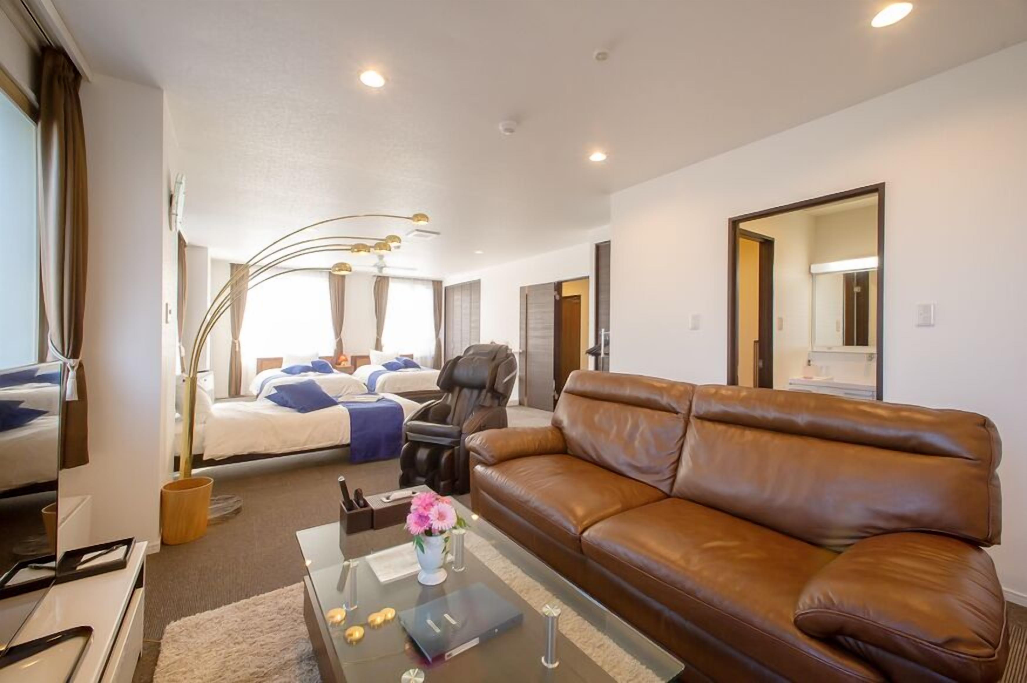 A modern hotel room with a bed, a leather sofa, a coffee table with a vase, and a desk with a computer.
