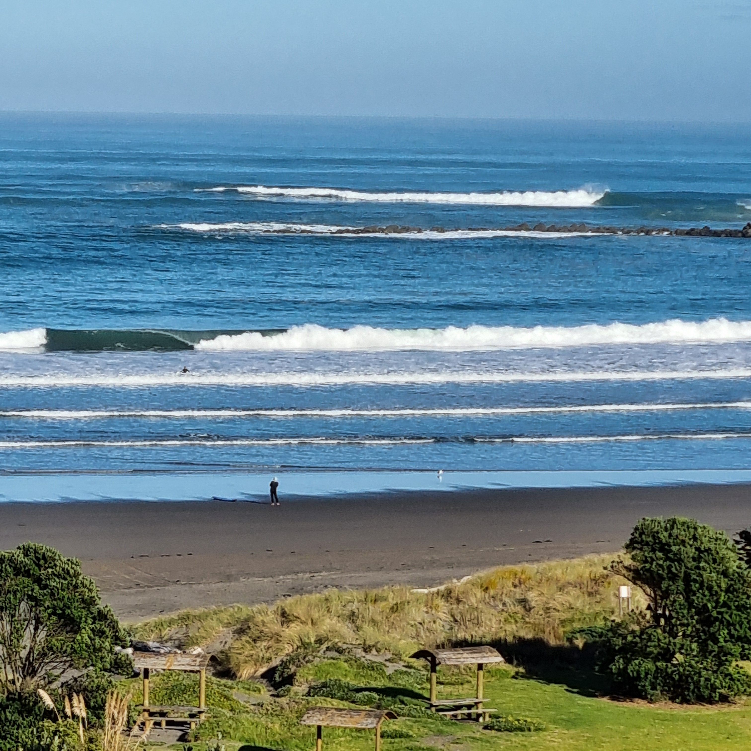 Une plage avec des vagues, une personne debout et quelques constructions au premier plan.