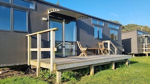 A modern mobile home with a deck, two wooden chairs, and a large sliding glass door. - Opunake Beach Kiwi Holiday Park (Opunake)