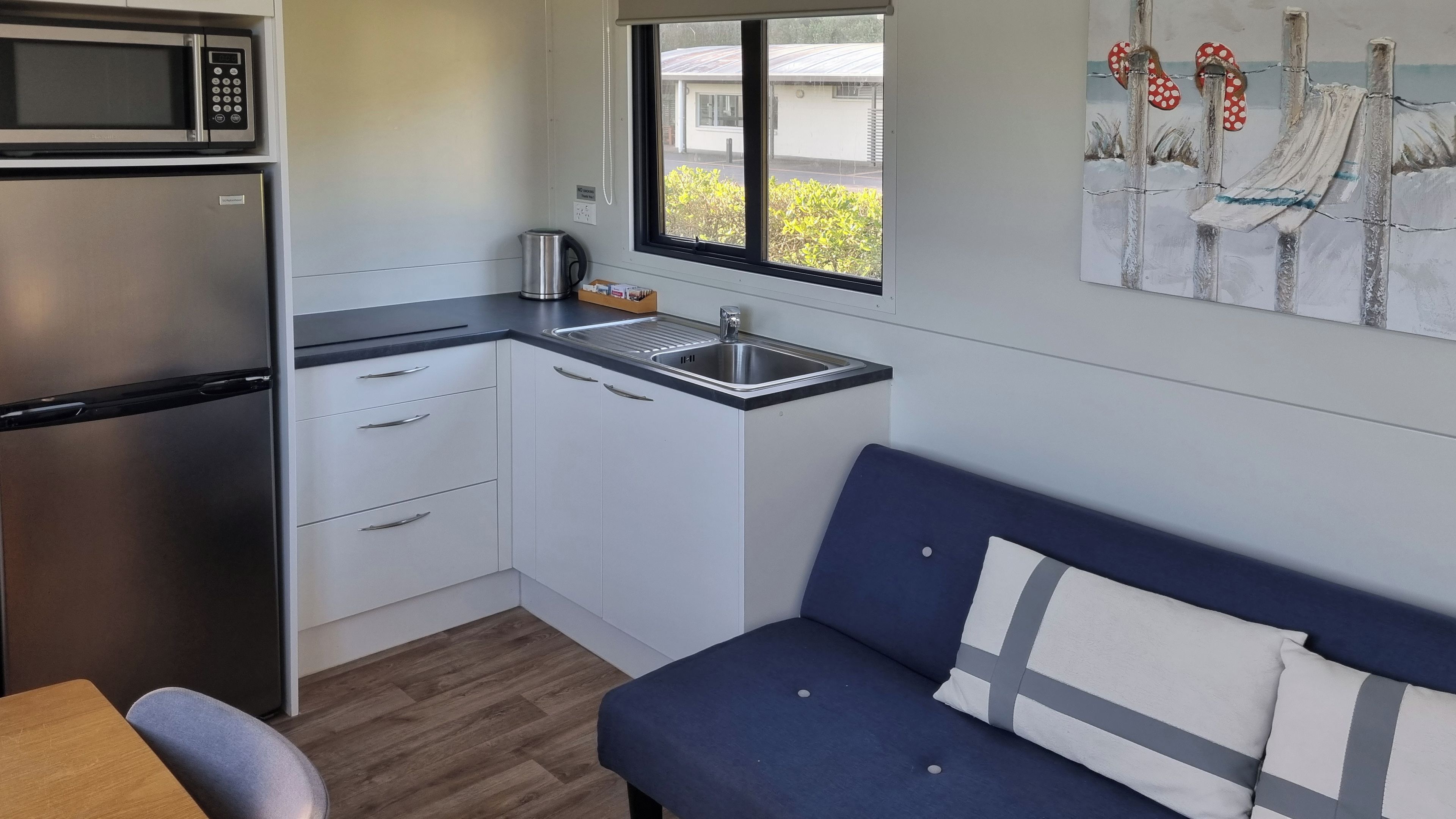 Cozinha compacta com geladeira de aço inoxidável, micro-ondas e pia. Há uma janela que oferece vista para um edifício e áreas verdes. Um sofá azul com uma almofada está encostado na parede, e uma mesa de madeira está em primeiro plano.