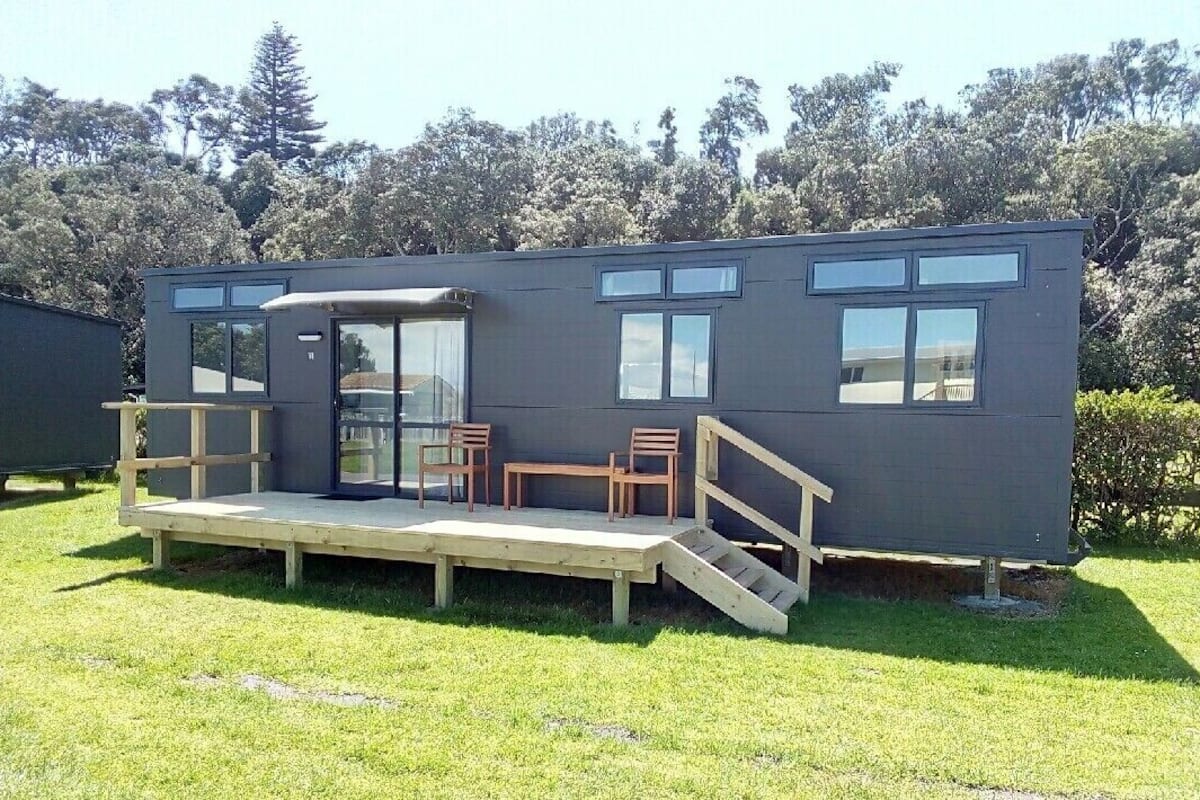A modern, compact mobile home with a small deck, two chairs, and a staircase leading to a door.