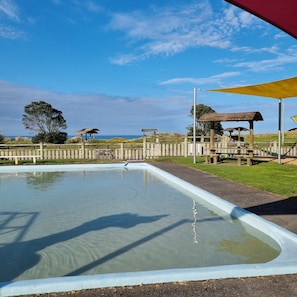 A swimming pool with a wooden deck and benches, surrounded by a wooden fence and trees. - Opunake Beach Kiwi Holiday Park (Opunake)