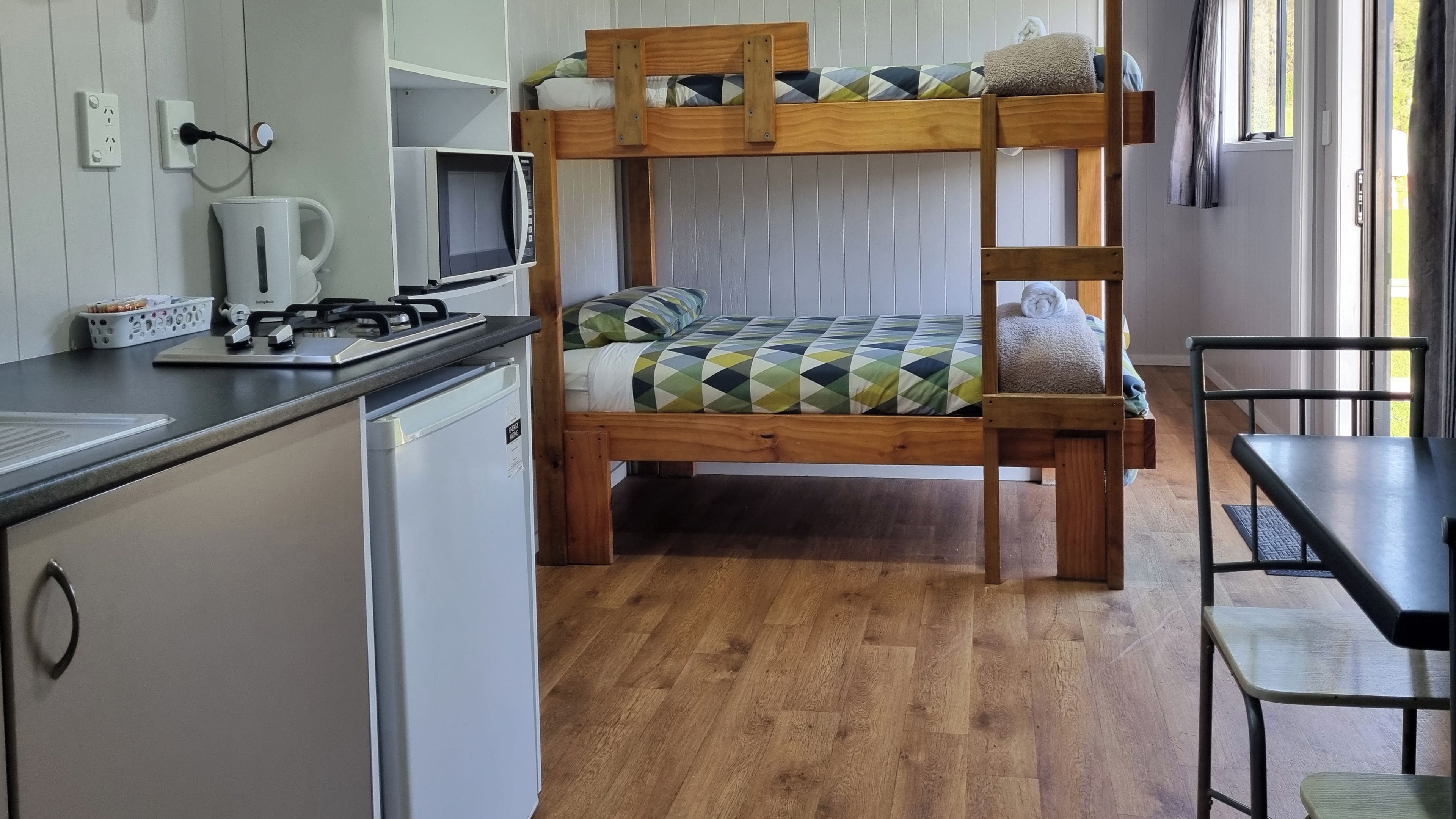 Een stapelbed met een geruit beddegoed, een kleine keuken met een fornuis en een spoelbak, en een raam met gordijnen.