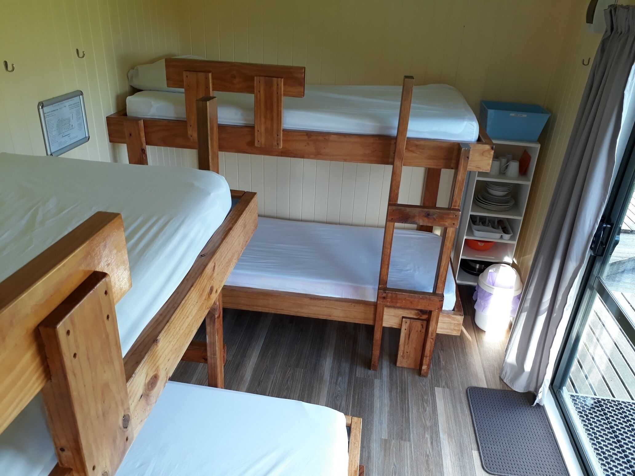 A bunk bed room with wooden frames, white bedding, a small shelf with dishes, and a window with curtains.
