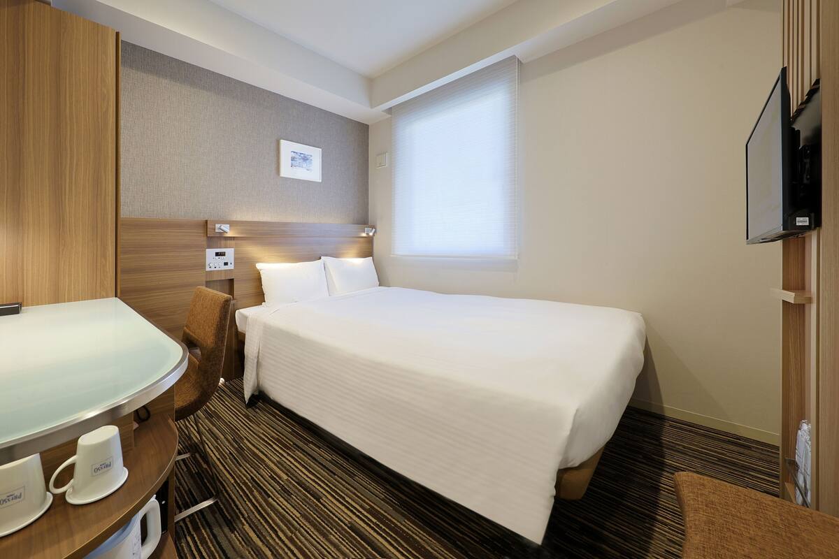 Una habitaci ón de hotel con cama, mesita de noche con taza, televisor y ventana.