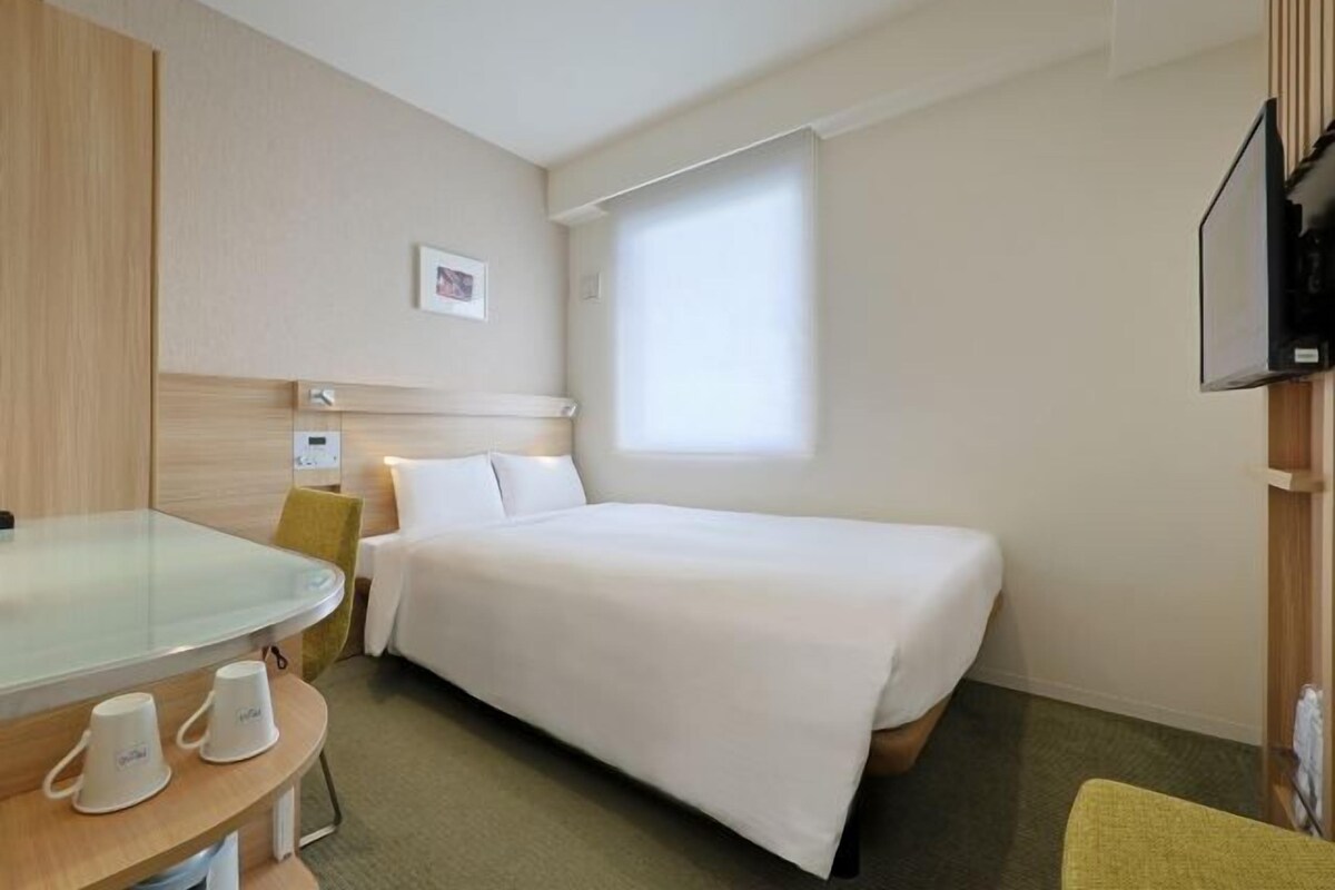 Habitación de hotel con cama, mesita de noche con una taza, una silla, un televisor y una ventana.