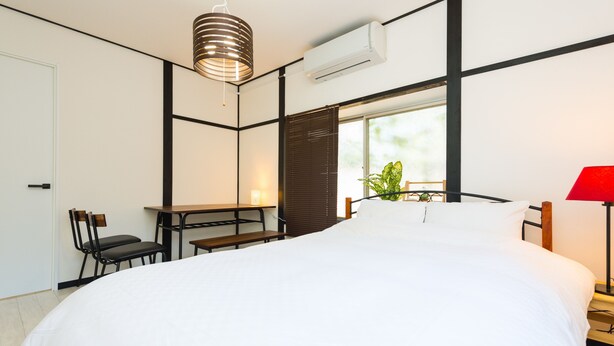 A modern bedroom with a large bed, a desk, chairs, a red lamp, and a window with blinds. - Otsu Ousaka House (Otsu)