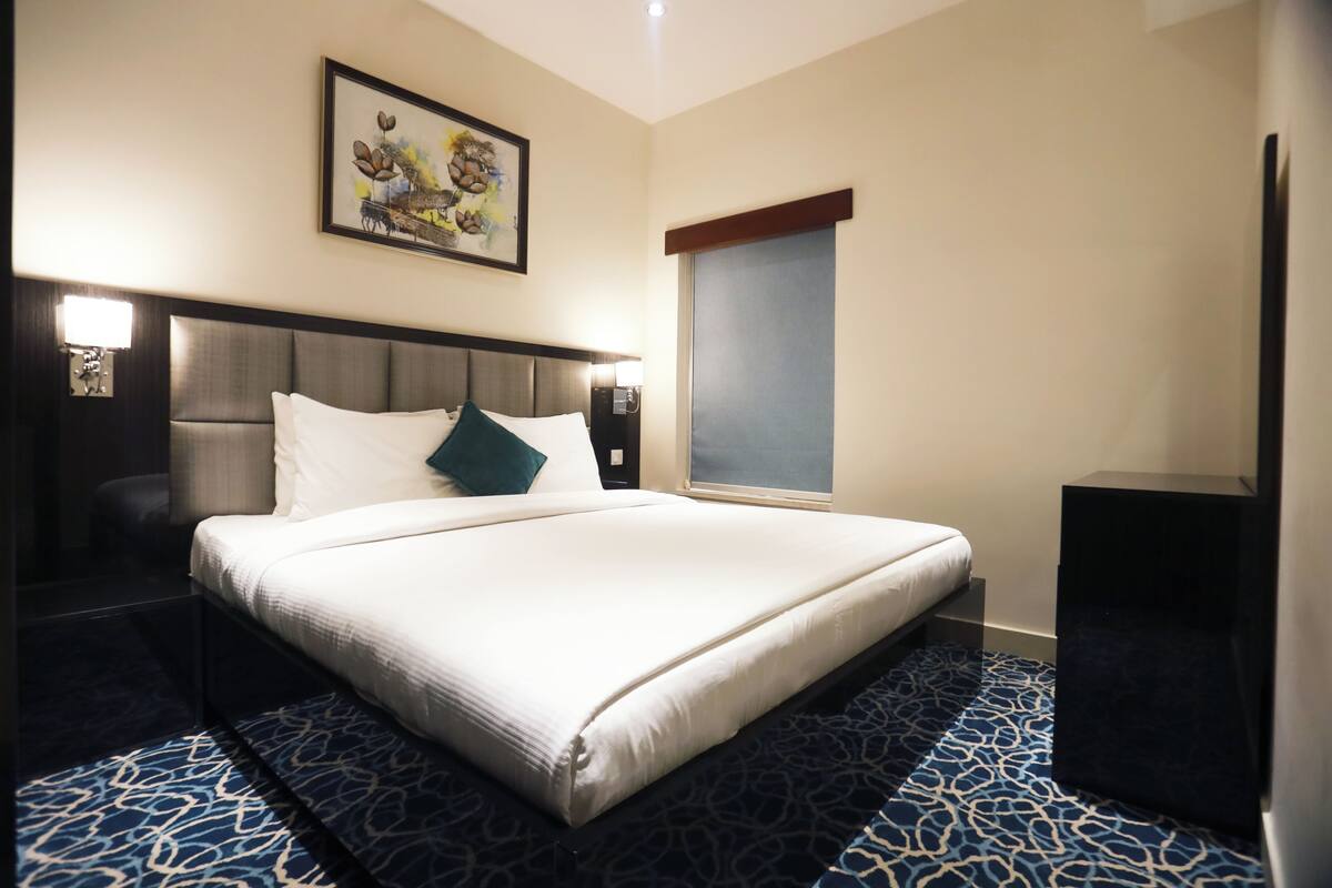 A hotel room with a large bed, a headboard, a nightstand, a wall-mounted lamp, a framed picture, a window with blinds, and a carpet with a blue and white pattern.