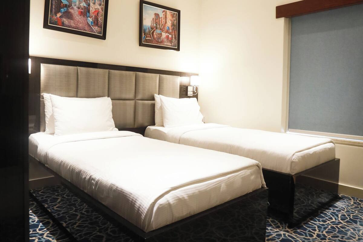 A hotel room with two beds, a headboard, a window, and two framed pictures on the wall.