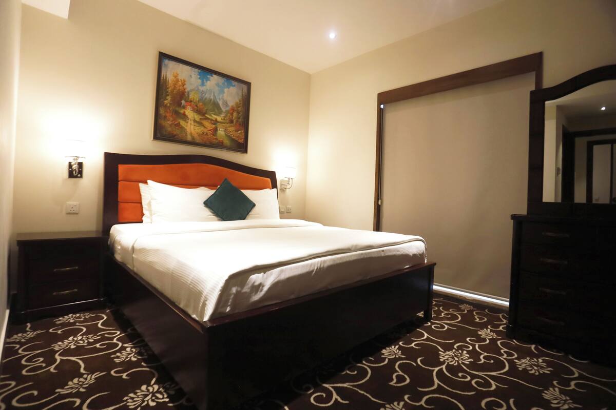 A bedroom with a large bed, a nightstand, a dresser, a painting on the wall, and a carpet with a floral pattern.