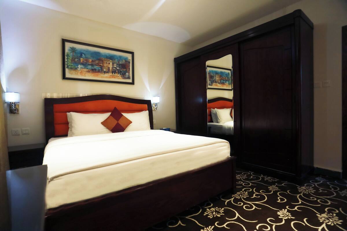 A hotel room with a large bed, a wooden wardrobe, a painting on the wall, and a patterned carpet.