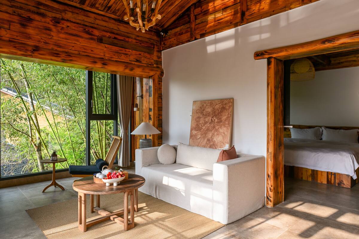 Een houten chalet van binnen met een bank, een bed, een tafel met fruit, en uitzicht op bomen door grote ramen.