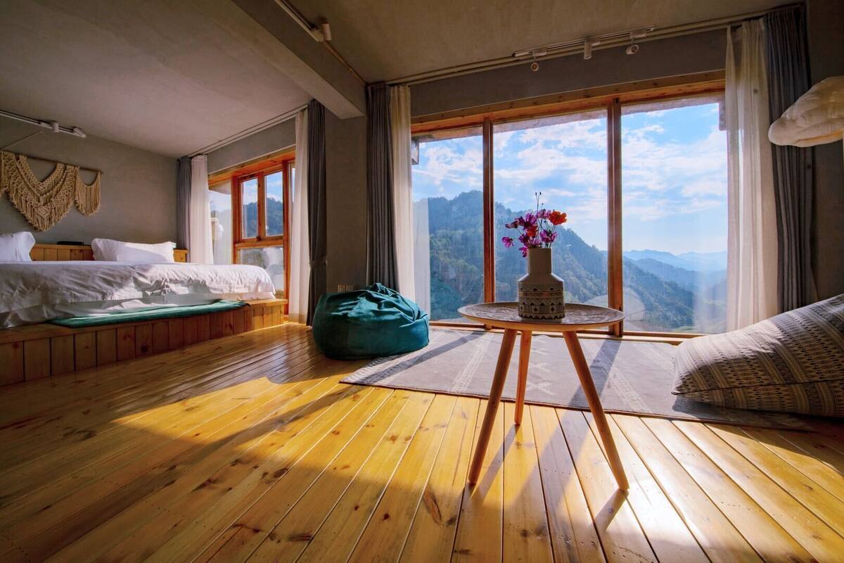 Een kamer met houten vloeren, een bed, een groene relaxfauteuil, een klein tafeltje met een bloevas en grote ramen met uitzicht op bergen.