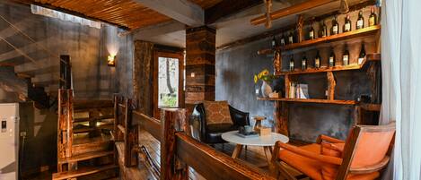 A rustic interior with wooden floors, a bar area with shelves of bottles, a seating area with a table and chairs, and a staircase.
