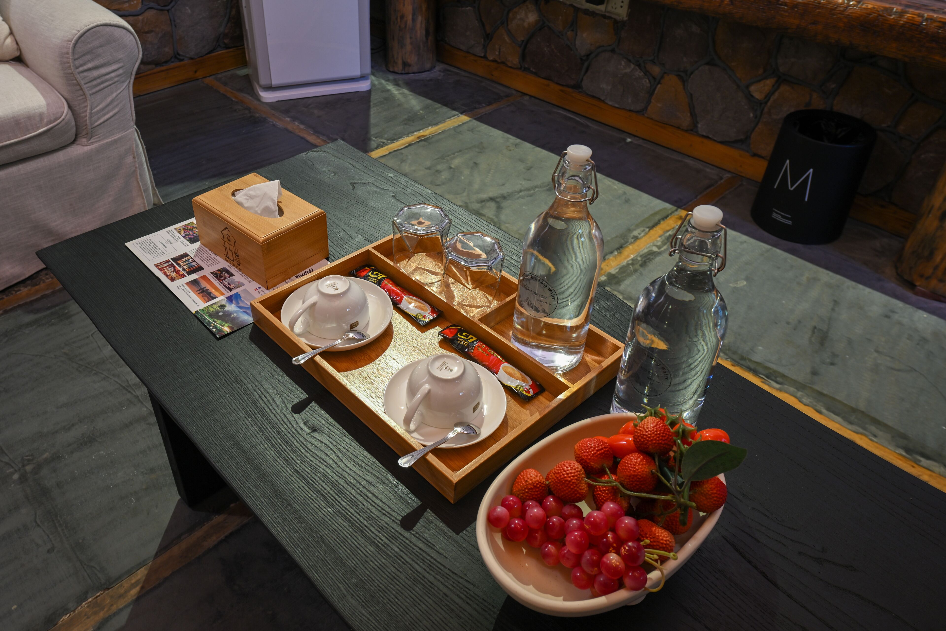 A breakfast setup with a tray, a bowl of strawberries, a bottle of water, and a tissue box on a wooden table.
