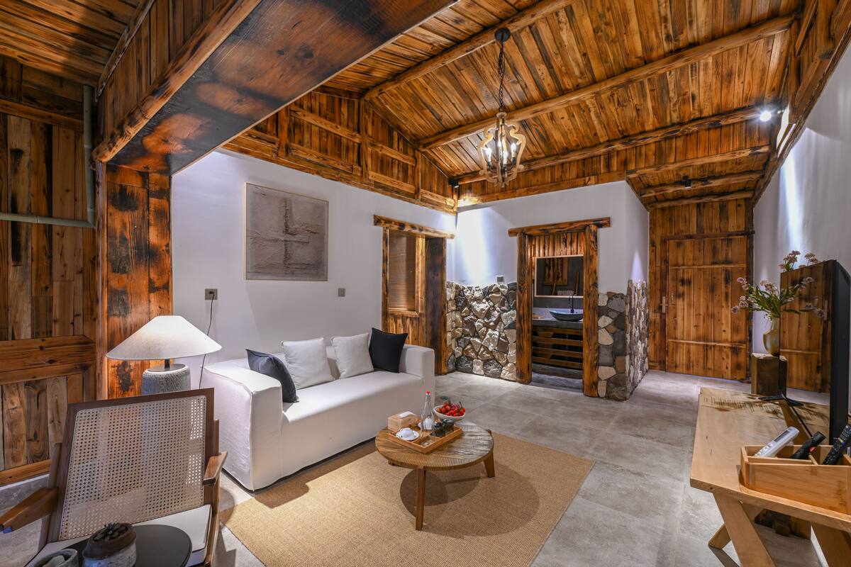 Een rustiek interieur met houten plafond, een witte bank, een stenen muur en een houten tafel met een vaas.