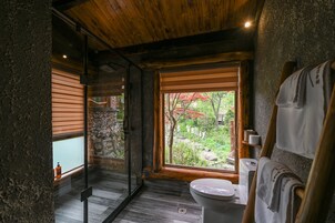 A bathroom with a wooden ceiling, a toilet, a sink, and a window with a view of a garden.