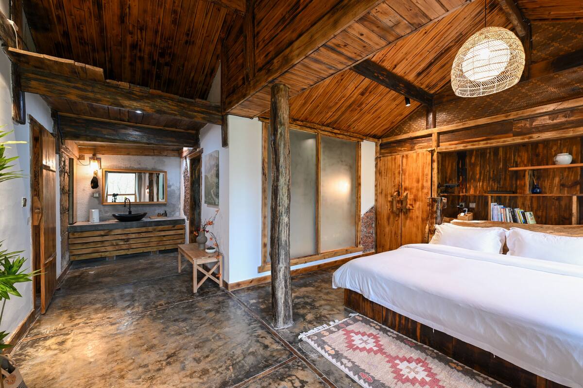 Een rustieke houten kamer met een groot bed, een houten bankje, een wastafel en een boekenplank.