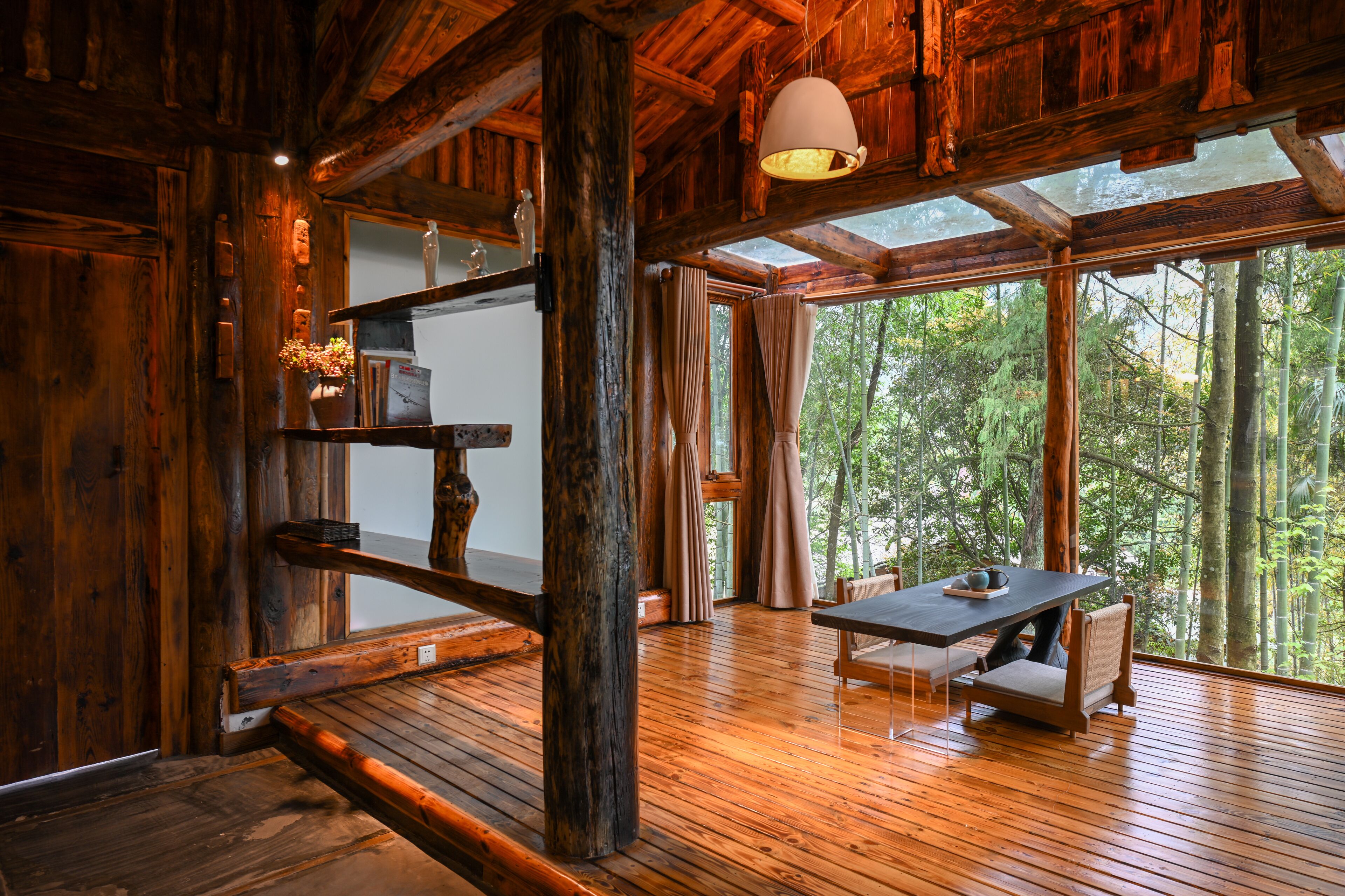 Un interior rústico de madera con una mesa y sillas de comedor, ventanas grandes que dan a un bosque y una lámpara colgante.
