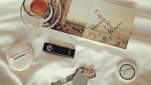 Una mesa con una taza de café, un libro titulado 'Capodimico Museo Real Botte', un bálsamo labial de Chanel, un llavero de B&B y un jabonera con la inscripción 'Mrs Smith'.