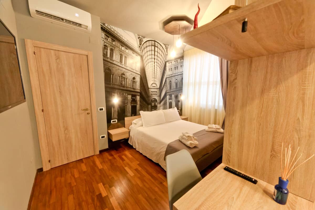 A hotel room with a bed, wooden floor, a desk with a vase, and a mural of a building on the wall.