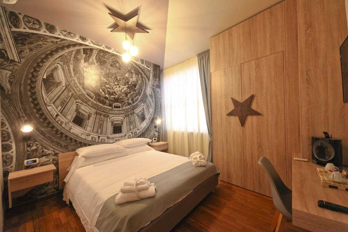 A hotel room with a large bed, a wooden wardrobe, a desk with a chair, a wall tapestry, and a ceiling light fixture.