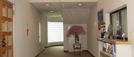 A hallway with a wooden door, a window with blinds, a vase with flowers, and a counter with various items.
