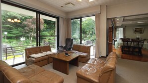 A spacious living room with leather sofas, a wooden coffee table, and large windows offering a view of a lush garden.