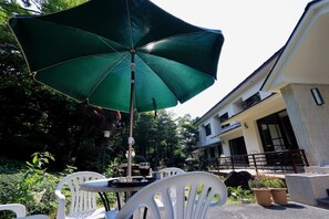 An outdoor patio with a table and chairs under a green umbrella, surrounded by potted plants and a building in the background. - Teiensaryo Yamanakako (Yamanakako)