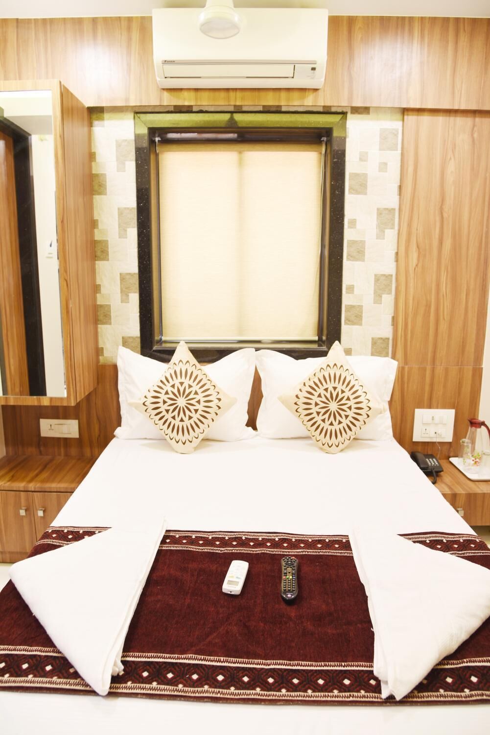 A hotel room with a bed, two decorative pillows, a remote control, and an air conditioning unit.