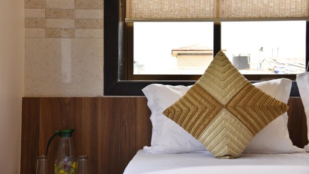 Habitación de hotel con cama, ventana que ofrece vista a edificios, cabecera de madera y mesita de noche con un tarro de vidrio y una botella verde.